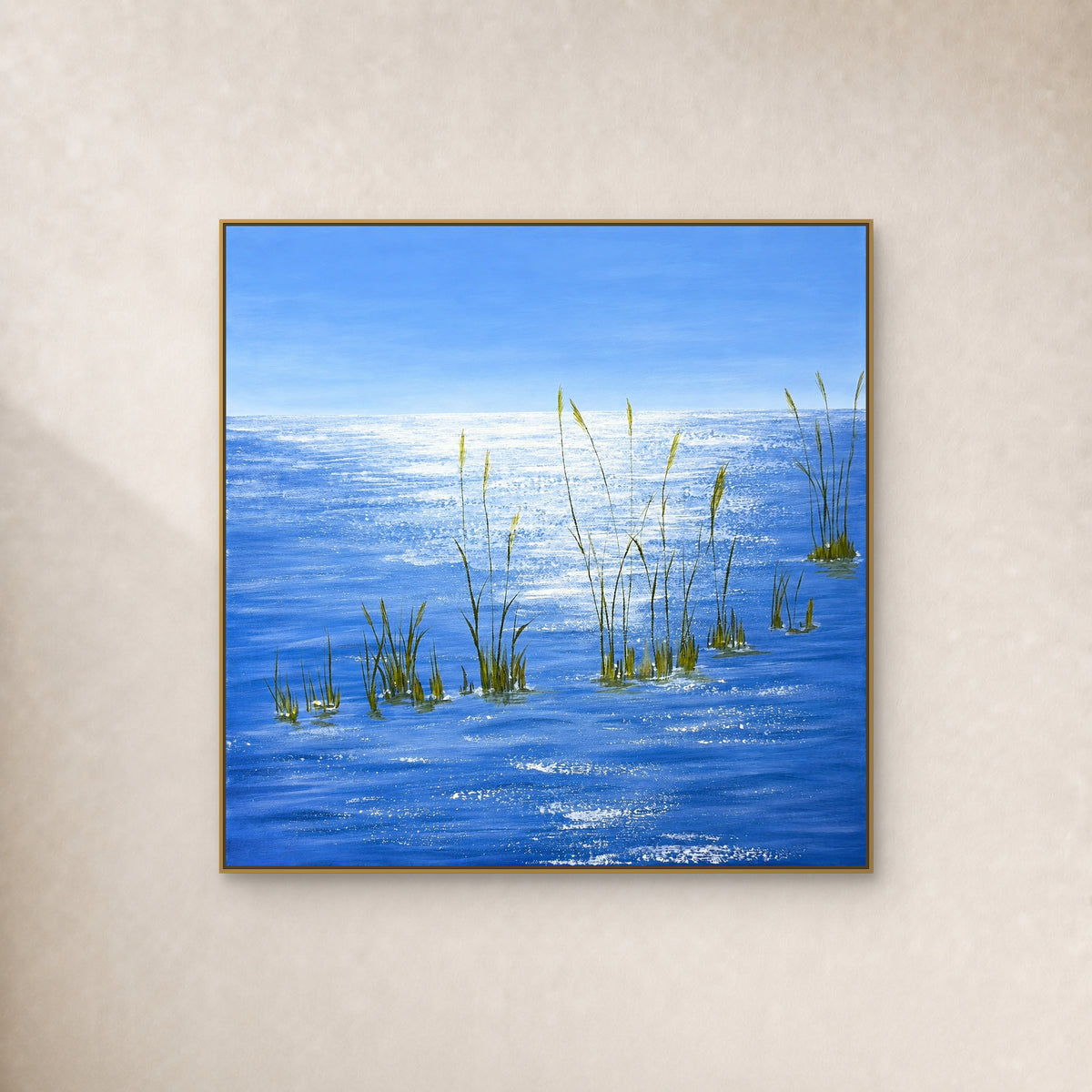 Tableau carré d’eau bleue, de reflets blancs et d’herbes vertes dans un fin cadre doré sur un mur ivoire