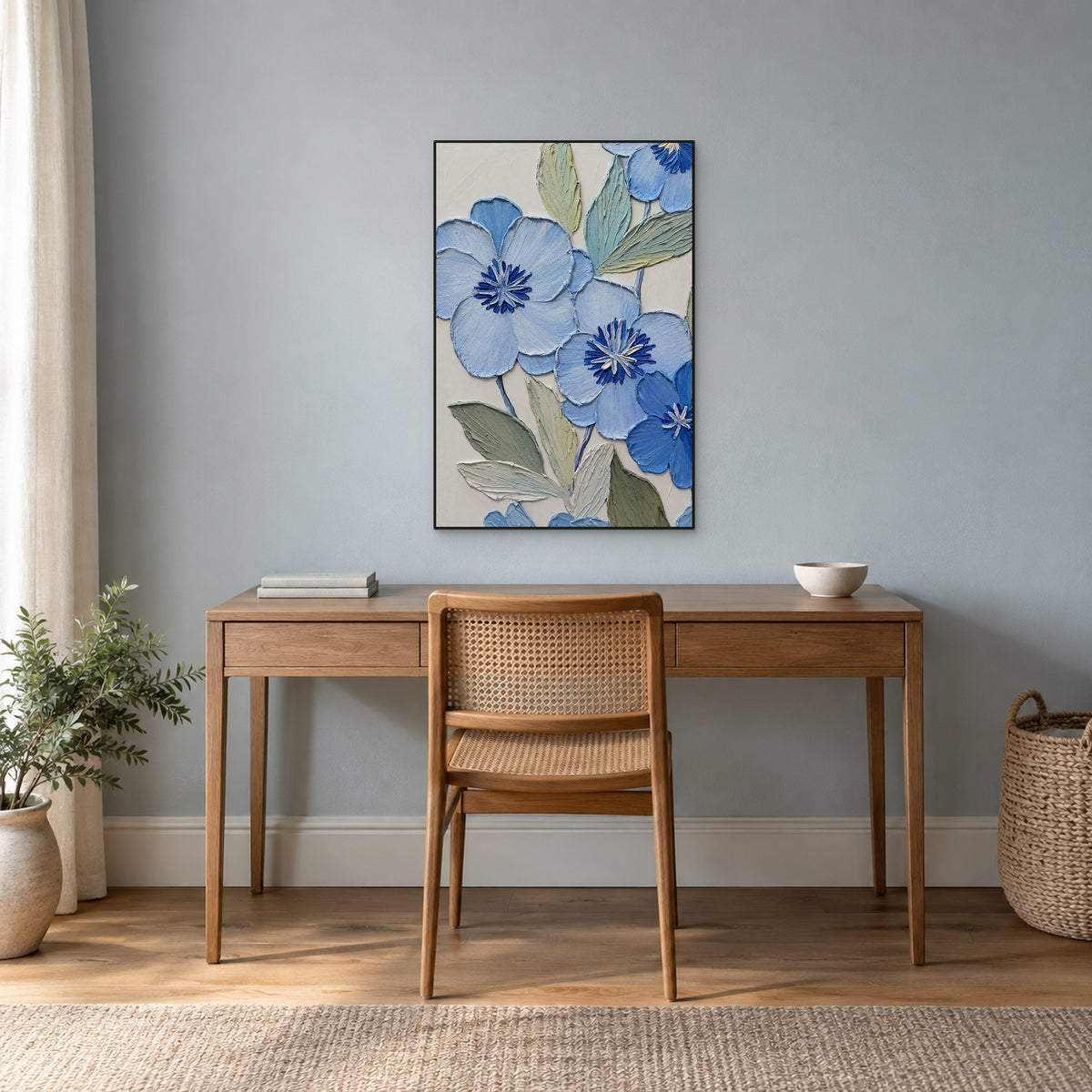 Blue flowers and sage leaves in a thin black frame above a wooden desk against a blue-grey wall