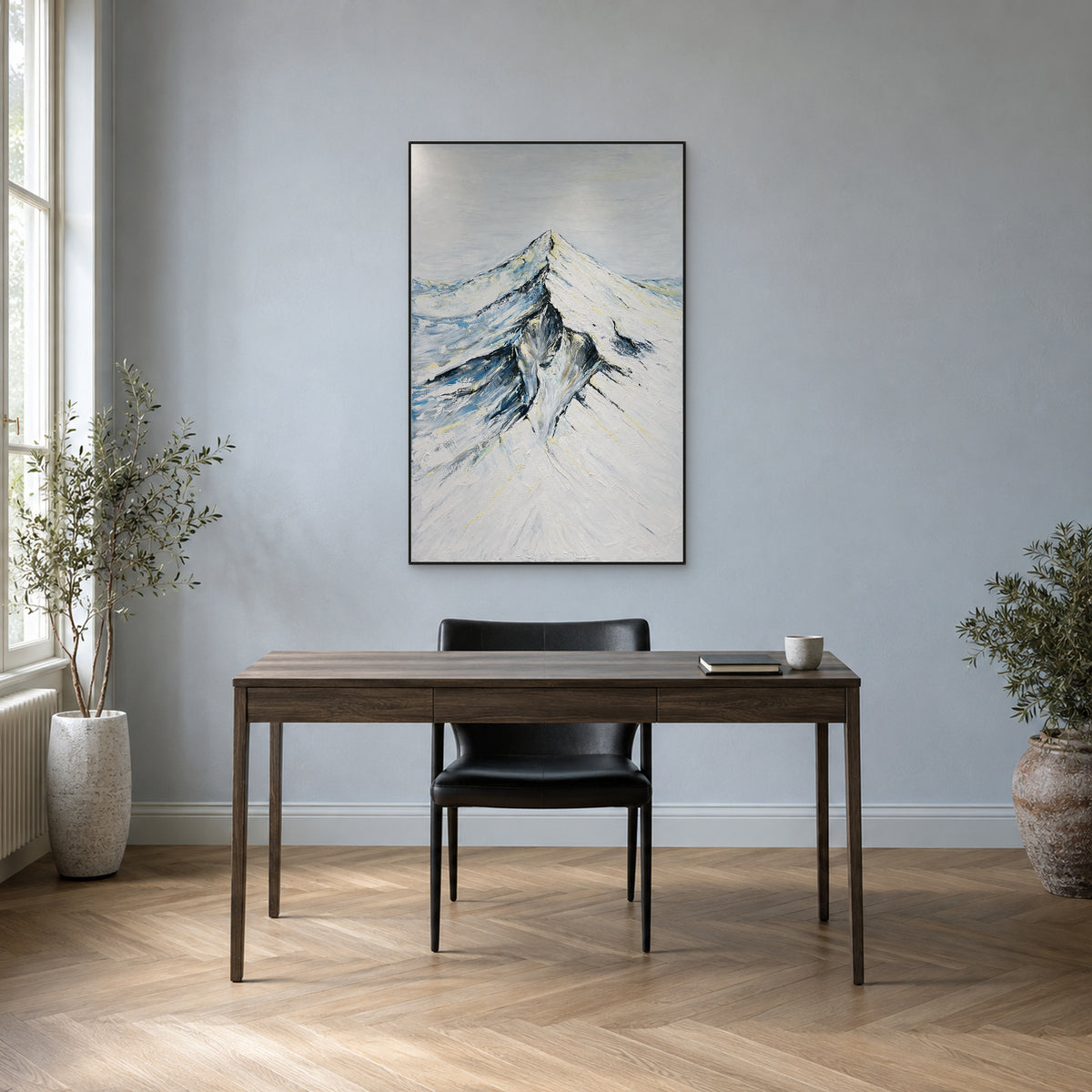 Snowy peak painting in a slim black frame above a walnut desk against a blue-grey wall