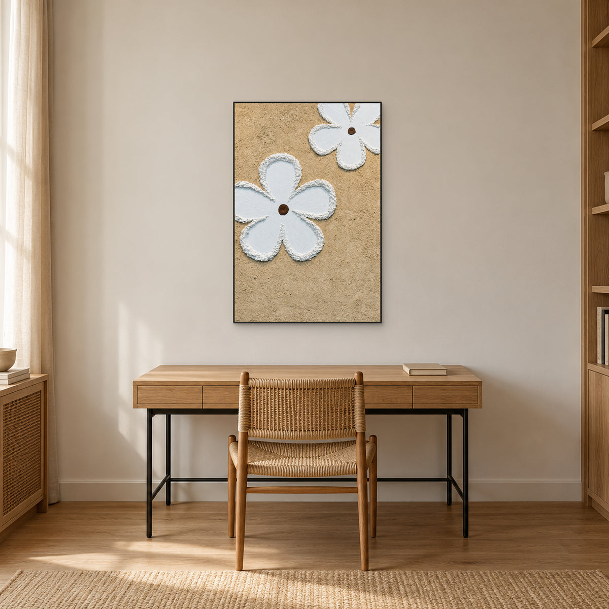 Beige and white flower painting in a thin black frame above a light wood desk and woven chair