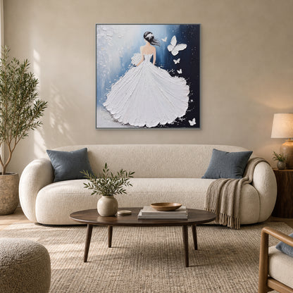 Square white gown painting with a silver-tone frame above a pale sofa with muted blue cushions
