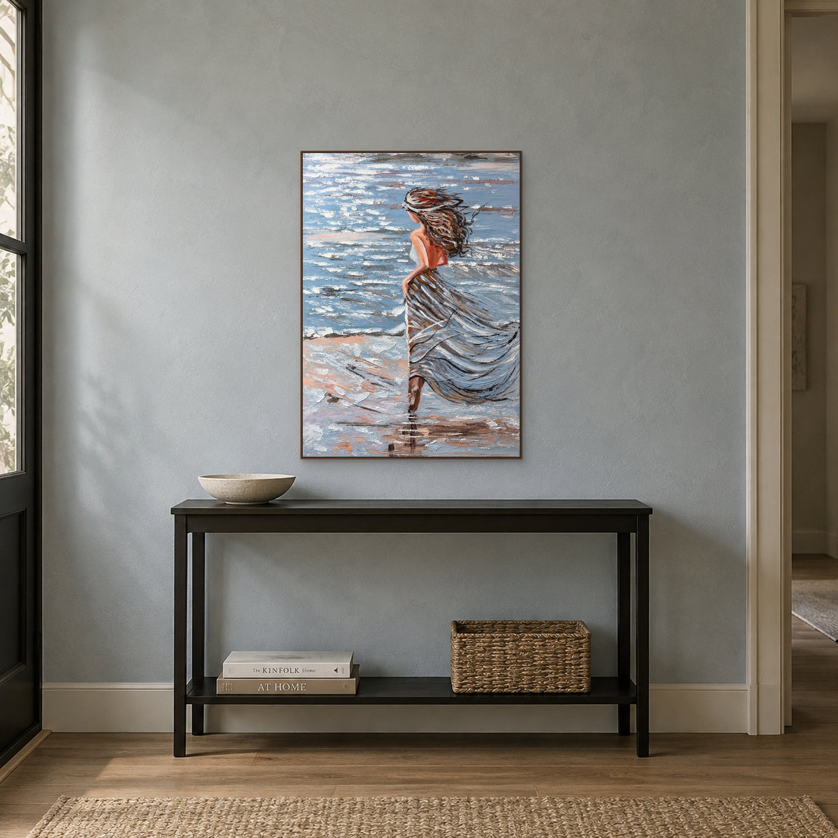 Tableau de femme au bord de mer dans un cadre flottant ton noyer, au-dessus d'une fine console noire devant un mur bleu grisé
