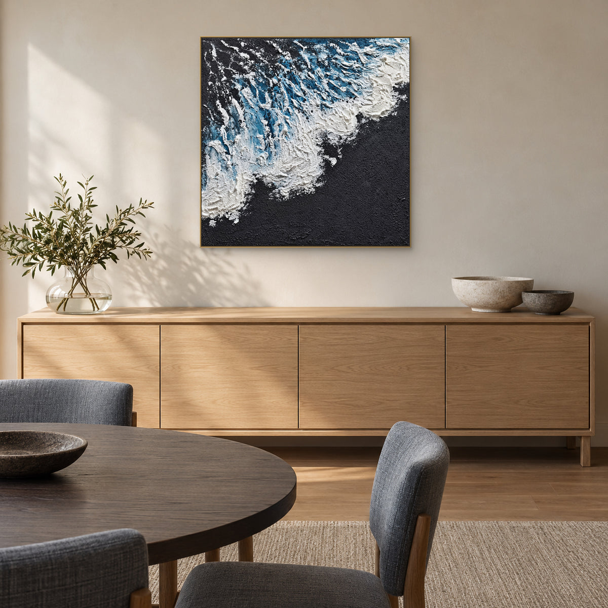 Square blue wave painting in a champagne-tone frame above an oak sideboard with a dining table in the foreground