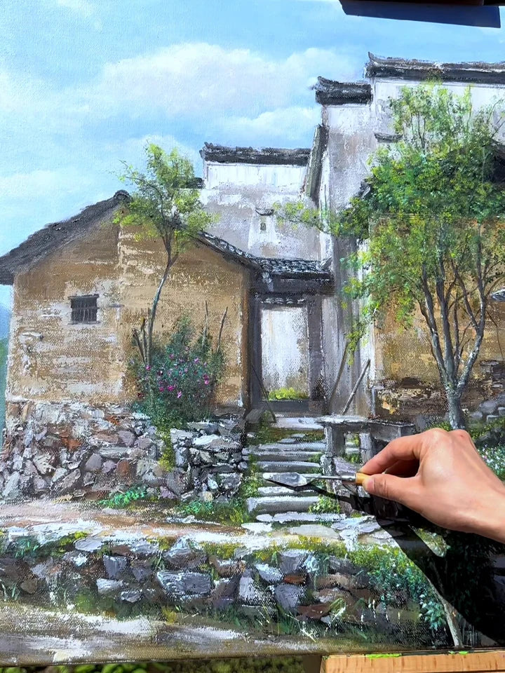 Un couteau à peindre ajoute des détails à un tableau de maisons de village traditionnelles et d’un chemin de pierre